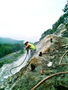 Saunders employee Josh Lucas using a rotary drill jack hammer drilling holes in ledge to later be blasted using explosives